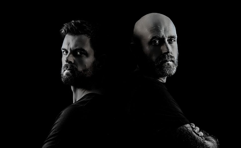 Metta and Glyde: two bearded men standing back to back against a black background, lit in dramatic black and white