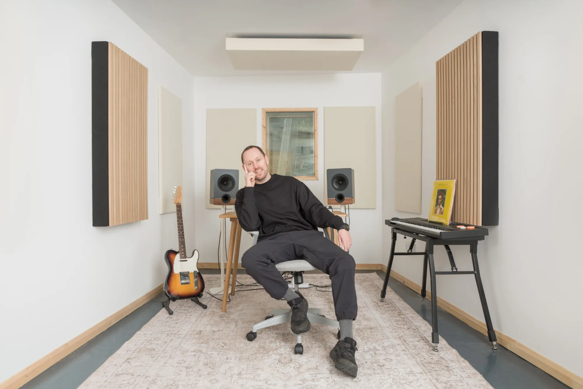 Will Evans seated in a bright studio room, with a guitar, studio speakers and a keyboard behind him